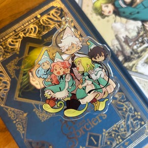 Può includere: Un ciondolo in acrilico trasparente con un gruppo di personaggi dei cartoni animati in abiti verdi e blu con cappelli a punta. Il ciondolo è su un libro blu con scritte dorate e il testo "Les Sorciers".