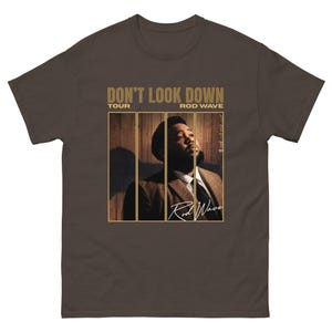 May include: A dark brown short-sleeve t-shirt featuring a graphic print of a person in a suit. The text DON'T LOOK DOWN TOUR ROD WAVE is printed in gold above the image, with the name Rod Wave in a white script font at the bottom right.