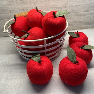May include: A white wire basket filled with several round, red felt apples, each featuring a small brown stem and a single green felt leaf. Three additional red felt apples with brown stems and green leaves are placed outside the basket.