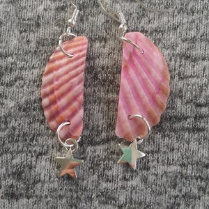 May include: A pair of pink seashell earrings with silver star charms. The earrings are made of natural seashells and have a pink and white color scheme.