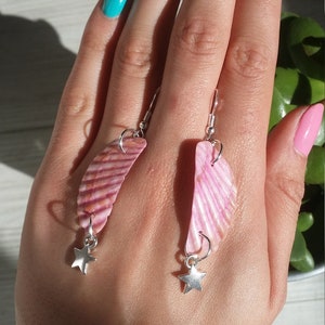 May include: A pair of pink shell earrings with silver star charms. The earrings are hanging from the earlobes of a person's hand.