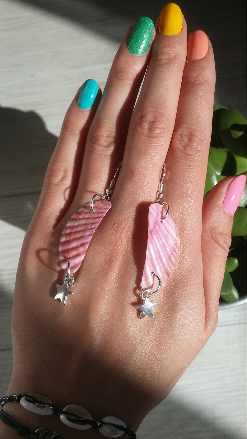 May include: A pair of pink shell earrings with silver star charms. The earrings are hanging from the earlobes of a person's hand.