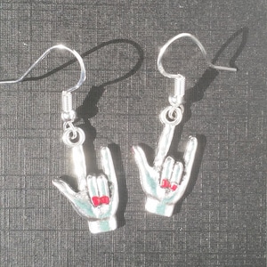 May include: A pair of silver-coloured earrings shaped like the ASL sign for "I love you." Each earring features a hand with a light blue enamel finish and red nail polish. The earrings hang from silver-coloured fish hook ear wires.