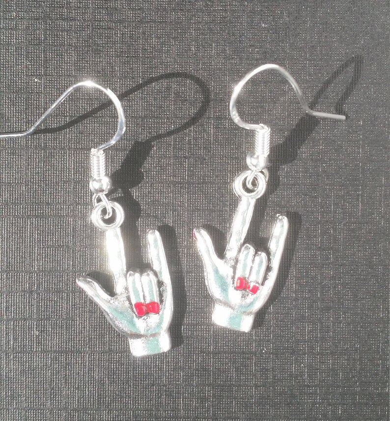 May include: A pair of silver-coloured earrings shaped like the ASL sign for "I love you." Each earring features a hand with a light blue enamel finish and red nail polish. The earrings hang from silver-coloured fish hook ear wires.