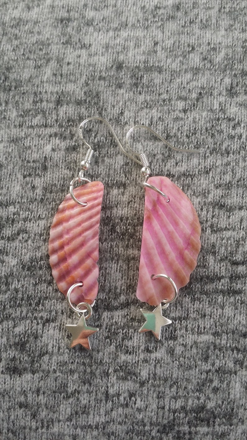 May include: A pair of pink seashell earrings with silver star charms. The earrings are made of natural seashells and have a pink and white color scheme.