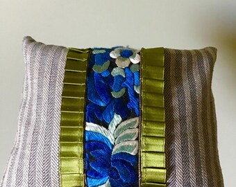 Könnte beinhalten: Kleines Zierkissen mit grau gestreiftem Stoff an den Seiten. Die Mitte zeigt eine lebendige blaue Blumenstickerei mit grünen und weißen Akzenten, umrahmt von grünem Band.