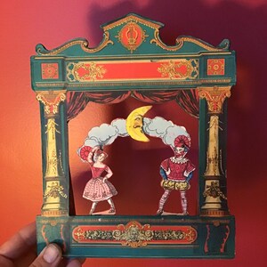 Peut inclure: Un théâtre de papier vintage avec un cadre vert et doré, des rideaux rouges et une lune souriante. Deux personnages en papier, une ballerine et un clown, sont sur scène. La scène est tenue par une main sur un fond rouge.