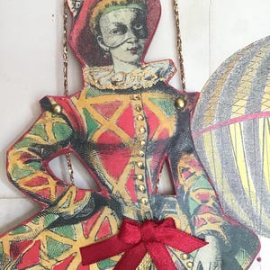 Peut inclure: Découpe en papier de style vintage représentant un personnage en costume d'arlequin. Le costume présente des motifs de losanges rouges, jaunes et verts. Le personnage porte un chapeau rouge, un col à volants et un masque.