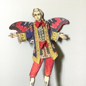 May include: A paper cutout of a figure in a historical costume with butterfly wings. The figure wears a blue jacket with gold trim, red pants, and a red bow tie. The butterfly wings are red and black.