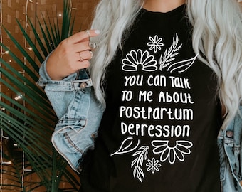 Może przedstawiać: Czarny t-shirt z białym rysunkiem liniowym kwiatów i liści otaczających napis "YOU CAN TALK TO ME ABOUT POSTPARTUM DEPRESSION" wykonany zaokrągloną czcionką. Koszulka jest noszona z niebieską kurtką dżinsową nałożoną na czarną warstwę bazową.
