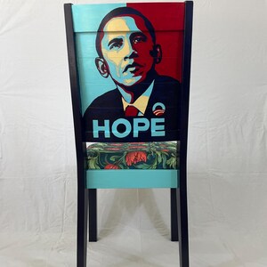 Puede incluir: Una silla de madera con un marco negro y un respaldo pintado de azul y rojo. El respaldo presenta un retrato de Barack Obama con la palabra "HOPE" impresa debajo. El asiento est&aacute; cubierto con una tela floral.