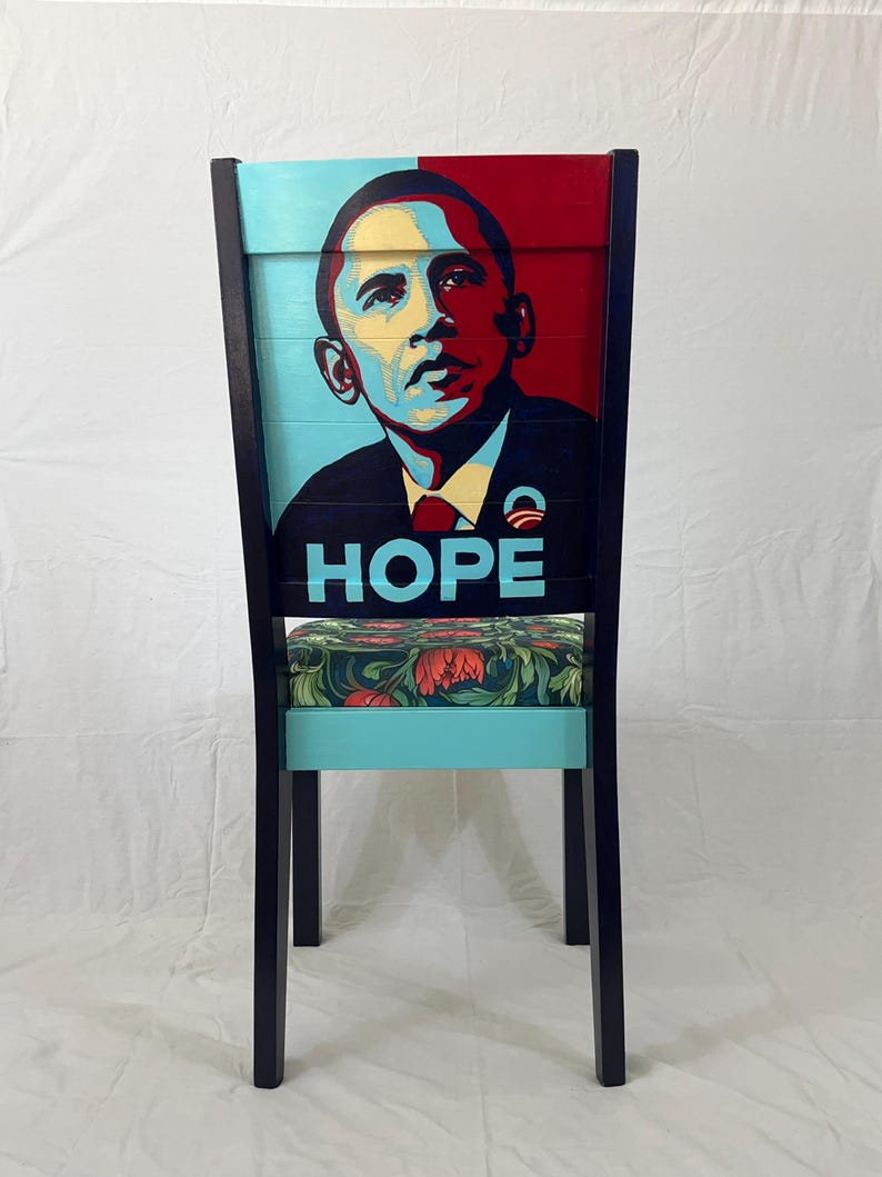 Puede incluir: Una silla de madera con un marco negro y un respaldo pintado de azul y rojo. El respaldo presenta un retrato de Barack Obama con la palabra "HOPE" impresa debajo. El asiento est&aacute; cubierto con una tela floral.