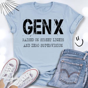 Puede incluir: Camiseta azul claro con el texto negro "GENX RAISED ON STREET LIGHTS AND ZERO SUPERVISION". La camiseta se combina con vaqueros azules y zapatillas blancas.