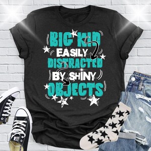 Puede incluir: Camiseta negra con el texto "BIG KID EASILY DISTRACTED BY SHINY OBJECTS" en turquesa y blanco. La imagen incluye zapatillas negras, vaqueros y calcetines con estampado de estrellas.