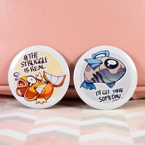 Puede incluir: Dos pines redondos con ilustraciones de peces de dibujos animados. El primer pin presenta un pez amarillo con el texto "#THE STRUGGLE IS REAL". El segundo pin presenta un pez azul y gris con el texto "I'LL GET THERE SOMEDAY..."