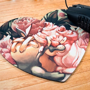 Puede incluir: Alfombrilla de ratón en forma de corazón con una ilustración caprichosa de un personaje de dibujos animados rodeado de flores rosas y melocotón. Un ratón de ordenador negro descansa en el lado derecho. El texto "@FRIEDUNICORNSTUDIO" es visible.