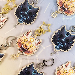 May include: Wooden wolf-head keychains in various colors, including black and tan, with gold-colored star and sun charms. The keychains have silver-colored clasps and are arranged on a light blue background with a constellation pattern. The text "@FRIEDUNICORNSTUDIO" is visible.