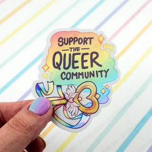 May include: A rainbow sticker with the text "Support the Queer Community" and a gold key with a heart-shaped hole and wings. The key is surrounded by a rainbow ribbon.