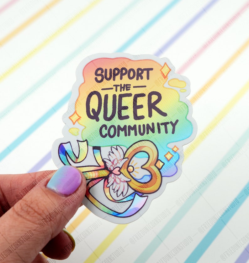 May include: A rainbow sticker with the text "Support the Queer Community" and a gold key with a heart-shaped hole and wings. The key is surrounded by a rainbow ribbon.