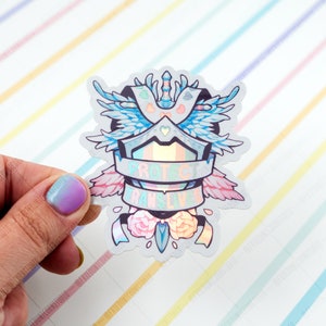May include: A holographic sticker featuring a shield, wings, and flowers. The shield has a rainbow stripe and the text "Protect Trans Lives".