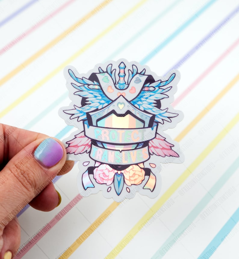 May include: A holographic sticker featuring a shield, wings, and flowers. The shield has a rainbow stripe and the text "Protect Trans Lives".