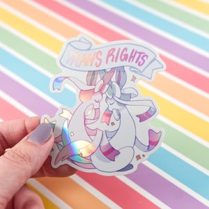 Puede incluir: Una pegatina holográfica con dos personajes de dibujos animados abrazándose. La pegatina tiene una pancarta que dice "TRANS RIGHTS" en texto blanco.