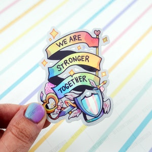 May include: A rainbow-coloured sticker with the words "We are stronger together" on three banners. The sticker also features a shield with a transgender pride flag, a key with wings, and a heart.