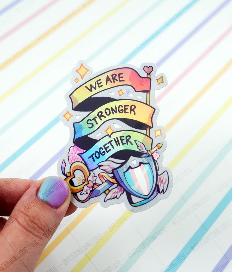 May include: A rainbow-coloured sticker with the words "We are stronger together" on three banners. The sticker also features a shield with a transgender pride flag, a key with wings, and a heart.