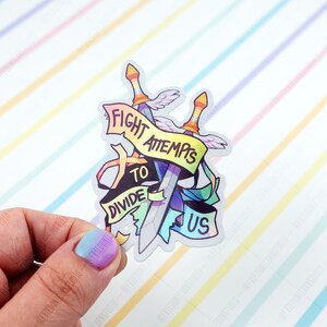 May include: A rainbow-coloured sticker with two swords crossed over each other. The swords are wrapped in a banner that reads "Fight Attempts to Divide Us".