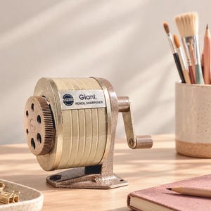 May include: A vintage Apsco Giant pencil sharpener with a beige cylindrical body, a brown metal base, and a hand crank. The front features a circular dial with multiple pencil holes and the text Giant PENCIL SHARPENER on a silver plate.