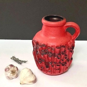 May include: A red ceramic vase with a textured, dark brown pattern. The vase has a handle and a dark brown interior. Two garlic bulbs and a sprig of dried herbs are next to the vase. The background is black and white.
