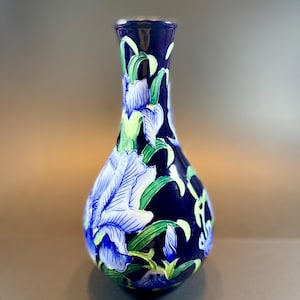 May include: A dark blue ceramic vase featuring a hand-painted floral design of light blue iris flowers with green leaves and stems. The vase has a narrow neck that widens into a rounded body, showcasing a glossy finish and detailed botanical art.
