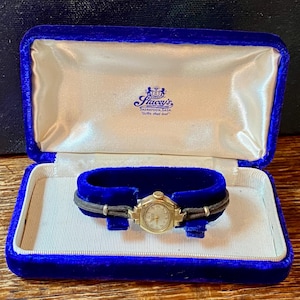 Peut inclure: Une montre-bracelet vintage dorée avec un petit cadran ovale, présentée dans un écrin en velours bleu. La montre est sur un bracelet en velours bleu avec un bracelet en cuir noir. La boîte est doublée de satin et porte le texte "Stacey's".