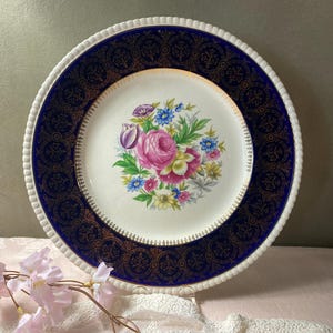 May include: A vintage porcelain plate with a white scalloped edge and a navy blue border with gold filigree. The center features a floral bouquet with pink roses and other colorful flowers. A decorative piece for serving.