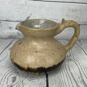 Może przedstawiać: Mały, ręcznie robiony ceramiczny dzbanek o zaokrąglonym korpusie i zakrzywionym uchwycie. Dzbanek ma nakrapiany, brązowy wygląd z ciemnobrązowym pasem u podstawy. Wnętrze jest jasnoszare. Dzióbek jest lekko spiczasty.