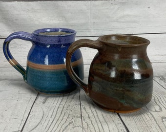 May include: Two handmade ceramic mugs. One mug is blue with brown and green accents. The other mug is brown with green and black accents. Both mugs have large handles and a rustic, handcrafted appearance.