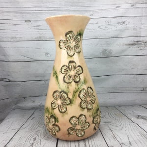 May include: A peach-colored ceramic vase with a flared top and a bulbous base. The vase is decorated with raised, textured floral designs in shades of brown and green. The vase is approximately 12 inches tall.