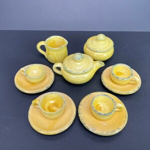 May include: A yellow ceramic tea set with a teapot, sugar bowl, creamer, and three teacups with saucers. The set has a rustic, handmade appearance with a speckled glaze and blue trim. The tea set is designed for a doll or child.
