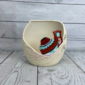 May include: Cream-colored ceramic bowl with a textured exterior and a brown, turquoise, and white decorative element. The bowl has a rounded shape with a cutout on one side, and the decorative element resembles a boot and a vase.