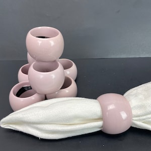 Può includere: Un set di sei anelli portatovaglioli in ceramica rosa pallido. Un anello tiene un tovagliolo di stoffa bianco. Gli anelli sono rotondi con una finitura liscia e lucida. Gli anelli portatovaglioli sono impilati a forma di piramide.