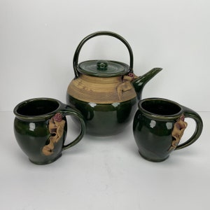 May include: A green and tan ceramic tea set. The set comprises a teapot with a handle and lid, and two matching mugs. Each piece features a decorative ribbon-like accent. The set is designed for serving tea.