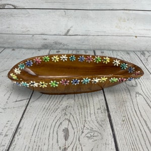 May include: A brown ceramic serving dish with a boat-like shape. The rim is decorated with colorful, raised floral accents in shades of blue, pink, yellow, green, and white. The dish has a glossy finish and is set against a light wood background.