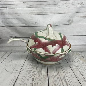 Puede incluir: Un cuenco de cerámica con tapa y cuchara adjunta. El cuenco es de color blanquecino con un patrón de pinceladas rojas y verdes. La tapa tiene un asa y la cuchara descansa en el lateral.