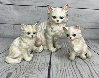 May include: Three white ceramic cat figurines. The largest cat is seated, with two smaller kittens flanking it. The cats have blue eyes and pink noses. The figurines are on a light-coloured wooden surface.