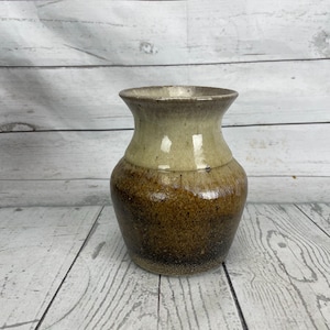 May include: A handcrafted ceramic vase with a speckled brown base that fades to a tan upper section. The vase has a wide, rounded body and a flared rim. The vase is approximately 6 inches tall.