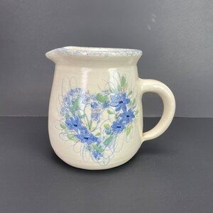 Puede incluir: Jarra de cerámica color crema con un diseño floral en forma de corazón en azul y verde. La jarra tiene un pico y un asa curva. El borde y el asa tienen un diseño moteado. El diseño floral está pintado a mano.