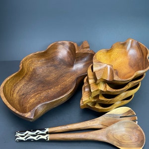 May include: A collection of wooden serving bowls and salad servers. The bowls are leaf-shaped and stacked, with a rich brown color. The salad servers have a light wood finish with decorative handles.