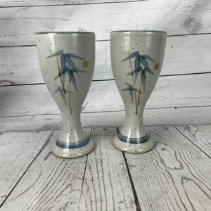 Puede incluir: Dos copas de cerámica con un acabado moteado blanquecino. Cada copa presenta un diseño de bambú azul y una banda azul cerca de la base. Las copas tienen un tallo delgado y una parte superior ancha y acampanada.