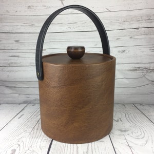 Puede incluir: Una cubitera cilíndrica marrón con asa y tapa negras. La tapa tiene un pequeño pomo redondo de madera. La cubitera tiene una superficie texturizada y un asa negra sujeta con un remache color latón.