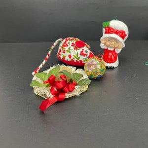 May include: A collection of Christmas ornaments, including a fabric mouse with a red floral pattern, a ceramic chef figurine, a decorative hat with a red bow, and a colorful round ornament. The ornaments are set against a dark background.